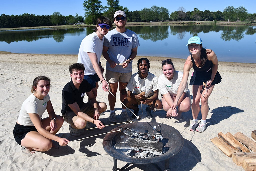 Students standing around a fire pit in front of a lake.