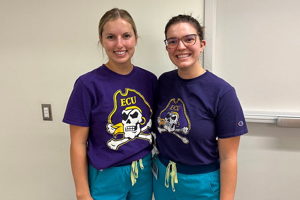 Two women with purple pirate shirts on in front of a wall.